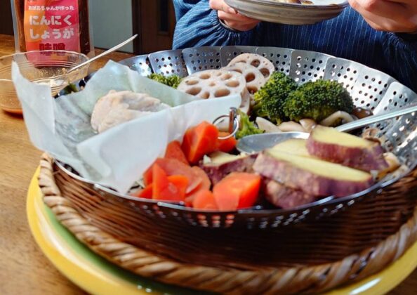 「蒸籠に盛りつけられた蒸し野菜と肉料理。さつまいも、にんじん、ブロッコリー、れんこん、きのこ類が並び、背景には箸と皿を持つ人物と『にんにくしょうが味噌』の瓶が見える。」