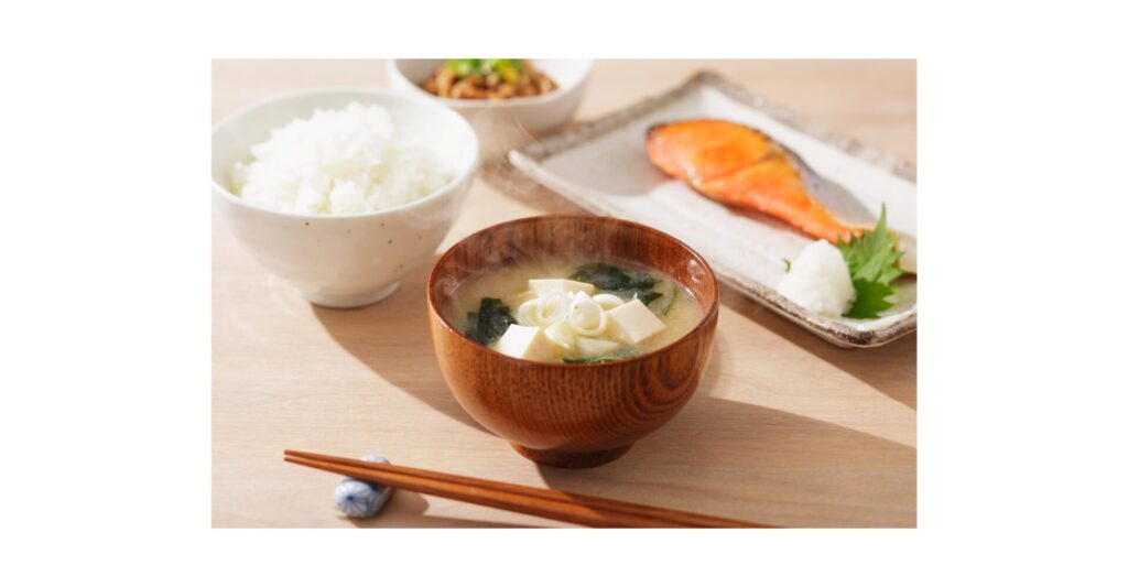 「木製のテーブルに並べられた和朝食セット。中央に味噌汁、左に白ごはん、奥に納豆、右に焼き鮭と大根おろし、手前には箸が置かれている。発酵食品を含む、栄養バランスのとれた日本の伝統的な朝食。」