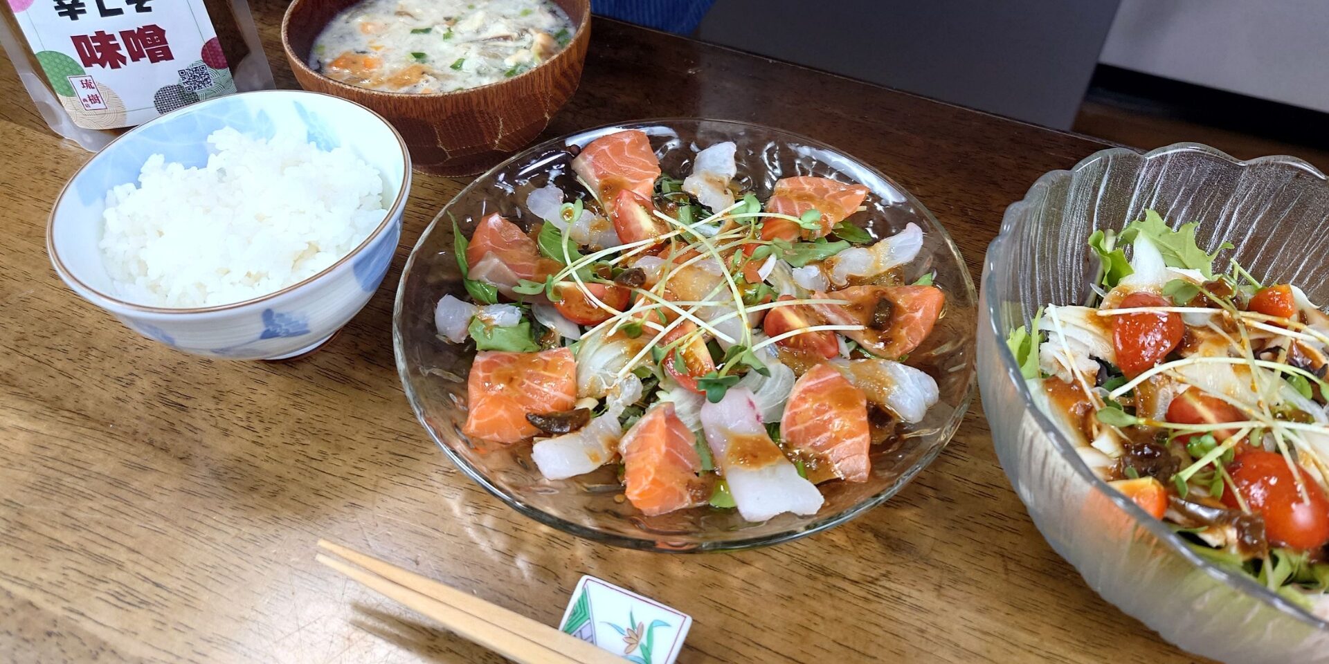 「木のテーブルに並べられた和朝食。白ごはん、豆腐とねぎ入りの味噌汁、サーモンと白身魚の刺身サラダ、彩り野菜のサラダ、辛口味噌の容器、箸と箸置きが整然と配置されている。」
