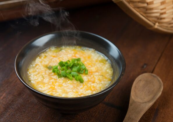 「木のテーブルに置かれた、湯気の立つ雑炊の器。ご飯と細かく刻まれた野菜が煮込まれ、優しい黄色のとろみが広がる。青ネギが彩りを添え、横には木製スプーンが添えられている。」