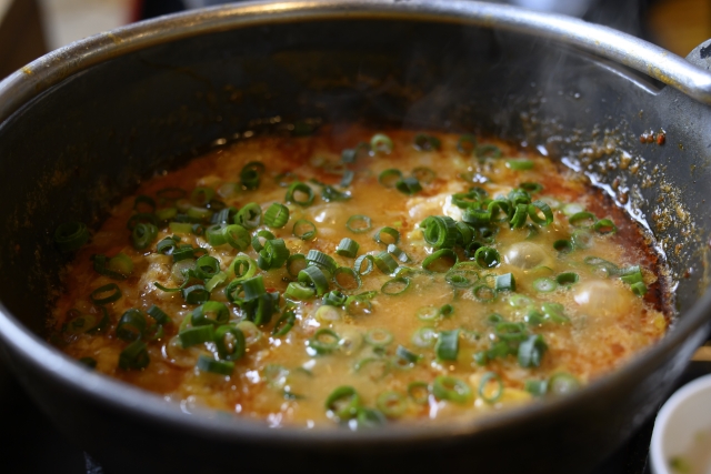 「赤いスープがグツグツと煮立つ鍋の中に、ご飯が加えられた雑炊。刻みネギが彩りを添え、旨味たっぷりの出汁がご飯に染み込んでいる様子が伝わる一枚。」