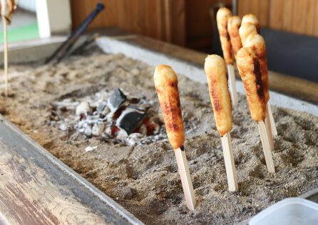「秋田の郷土料理・きりたんぽの原型。炭火を囲む砂床で串焼きされたご飯棒が、伝統的な調理法で香ばしく焼き上げられる様子。」