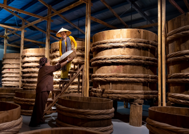 「木桶が並ぶ味噌蔵で、冬の寒仕込み作業を行う職人たち。ひとりが梯子に登り、もうひとりが材料を手渡す様子が映し出され、伝統的な手仕事の温もりと丁寧な工程が感じられる。」